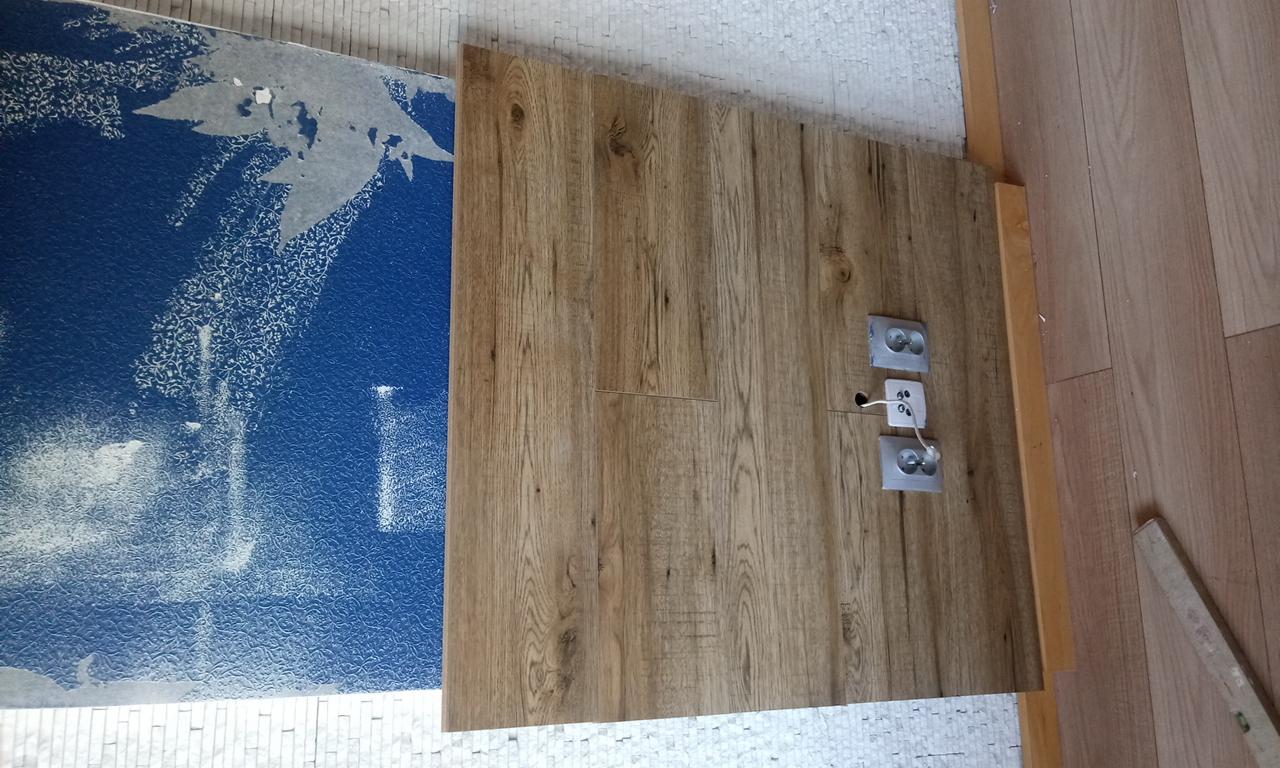 Fragment podłogi z paneli drewnopodobnych w trakcie montażu, widoczne gniazdka elektryczne, niebieska folia zabezpieczająca i fragment istniejącej podłogi.