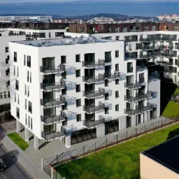 Nowoczesny, biały budynek mieszkalny z balkonami z metalowymi balustradami, widziany z lotu ptaka, z zielonym trawnikiem i drzewami w tle.