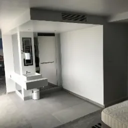 Nowoczesna łazienka połączona z sypialnią, z przeszkloną kabiną prysznicową, minimalistyczną umywalką na blacie i szarą podłogą z płytek, widok na część sypialnianą z tapetą z motywem pejzażu.