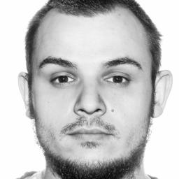 Marcin Gregorek - Czarno-biały portret mężczyzny z krótkimi włosami, brodą i wąsami, na białym tle. Widoczny pieprzyk na policzku.