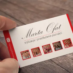 Dłoń trzyma wizytówkę z imieniem Marta Ofat, opisem stylizacja i przedłużanie paznokci oraz miniaturami zdjęć paznokci na drewnianym tle.
