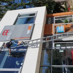 Mężczyzna na drabinie myje okna biurowca z logo 'Autoreflex' i 'ProfiAuto.pl', widoczne narzędzia i szyld reklamowy.