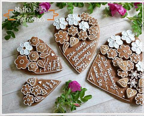 Cztery piernikowe serca w różnych rozmiarach, zdobione białymi lukrowanymi kwiatami i dedykacjami pisanymi lukrem, ułożone na jasnym drewnianym tle, obok gałązka różowych róż.