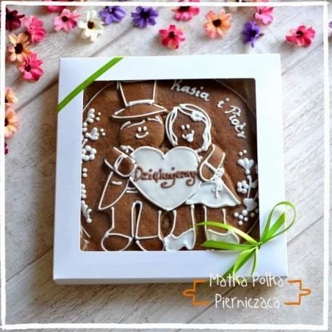 Piernikowe ciasteczko w pudełku z motywem pary młodej, dekorowane lukrem, z napisem 'Dziękujemy' i imionami Kasia i Piotr, otoczone kwiatami.