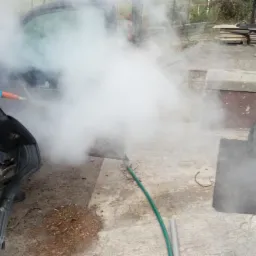 Mężczyzna w zielonej bluzie i niebieskich rękawicach parowo czyści wnętrze samochodu, obok leżą wyjęte dywaniki, kłęby pary unoszą się w powietrzu.