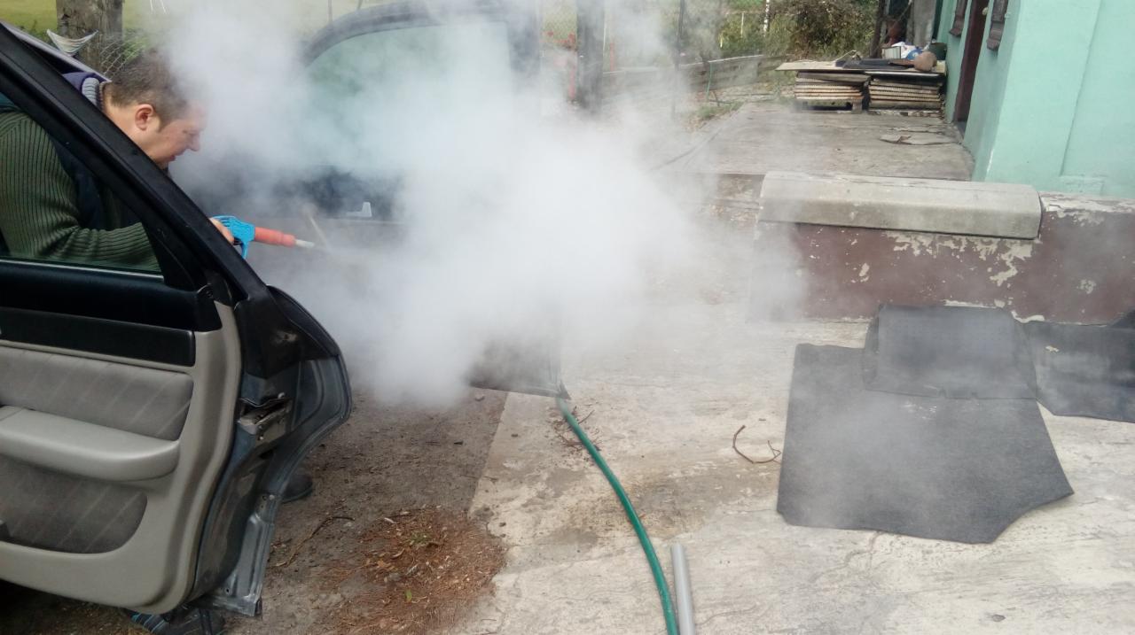 Mężczyzna w zielonej bluzie i niebieskich rękawicach parowo czyści wnętrze samochodu, obok leżą wyjęte dywaniki, kłęby pary unoszą się w powietrzu.