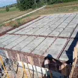 Wieniec stropowy z szalunkiem i zbrojeniem, widoczne podpory stropowe i elementy prefabrykowane w tle krajobraz wiejski.