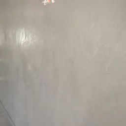 Ściana w łazience wykończona dekoracyjną, szarą powłoką imitującą beton, z widoczną fakturą i połyskiem, oświetlona światłem padającym z lewej strony.