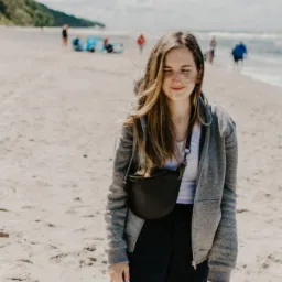 Uśmiechnięta młoda kobieta na plaży, w szarej bluzie i z czarną torebką na biodrach, w tle morze i ludzie. Włosy rozwiane na wietrze.