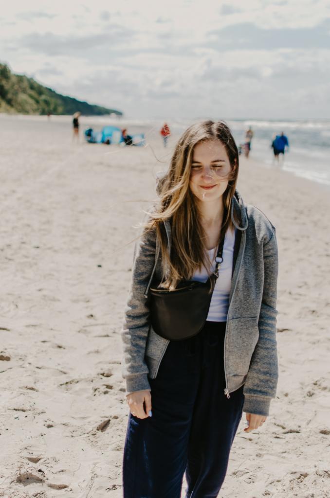 Uśmiechnięta młoda kobieta na plaży, w szarej bluzie i z czarną torebką na biodrach, w tle morze i ludzie. Włosy rozwiane na wietrze.