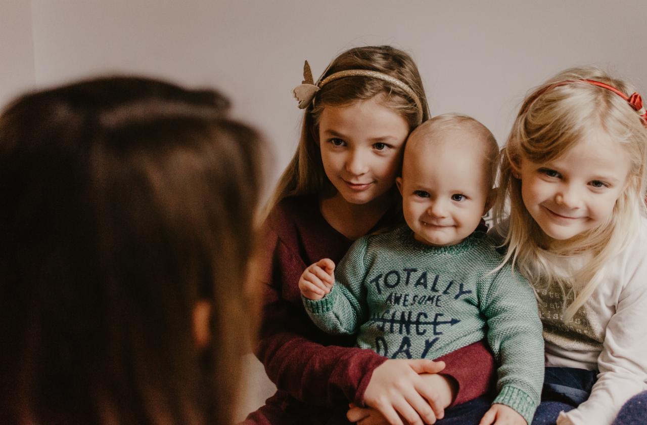 Uśmiechnięte rodzeństwo: starsze siostry obejmują młodszego brata w zielonym swetrze z napisem, ciepłe, naturalne światło.