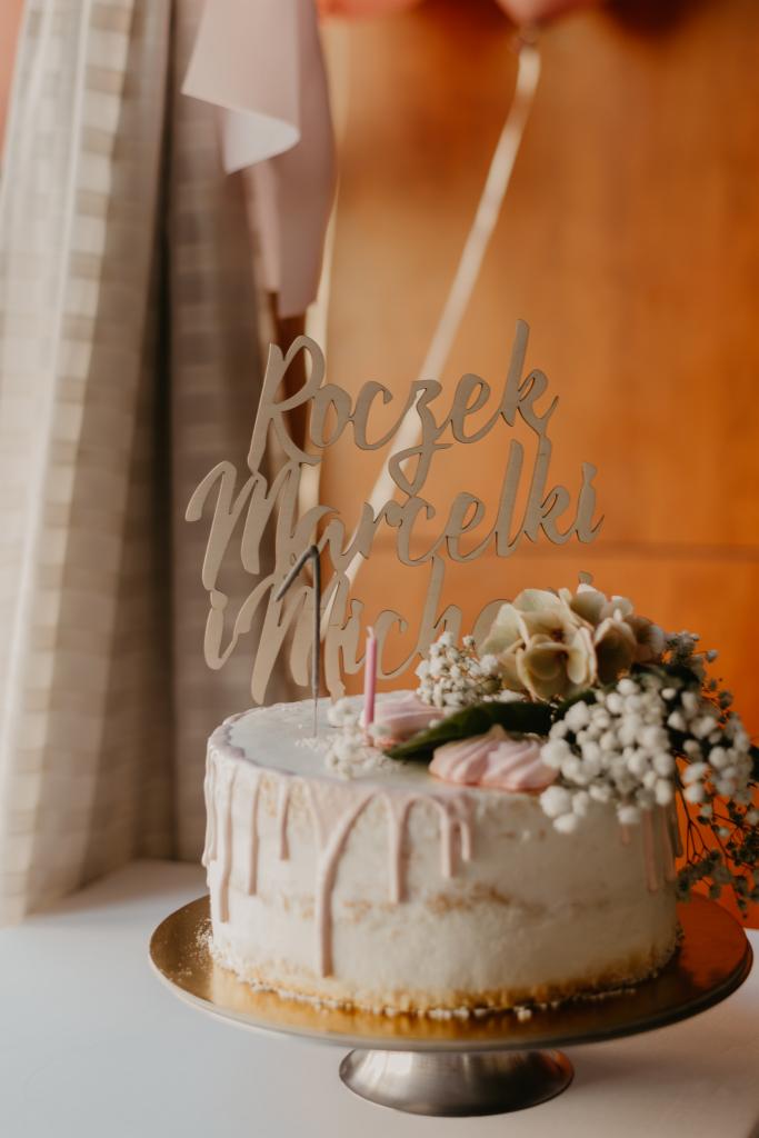 Tort z napisem 'Roczek Marcelki i Michała', udekorowany kwiatami i różowymi bezami, na złotym talerzu.