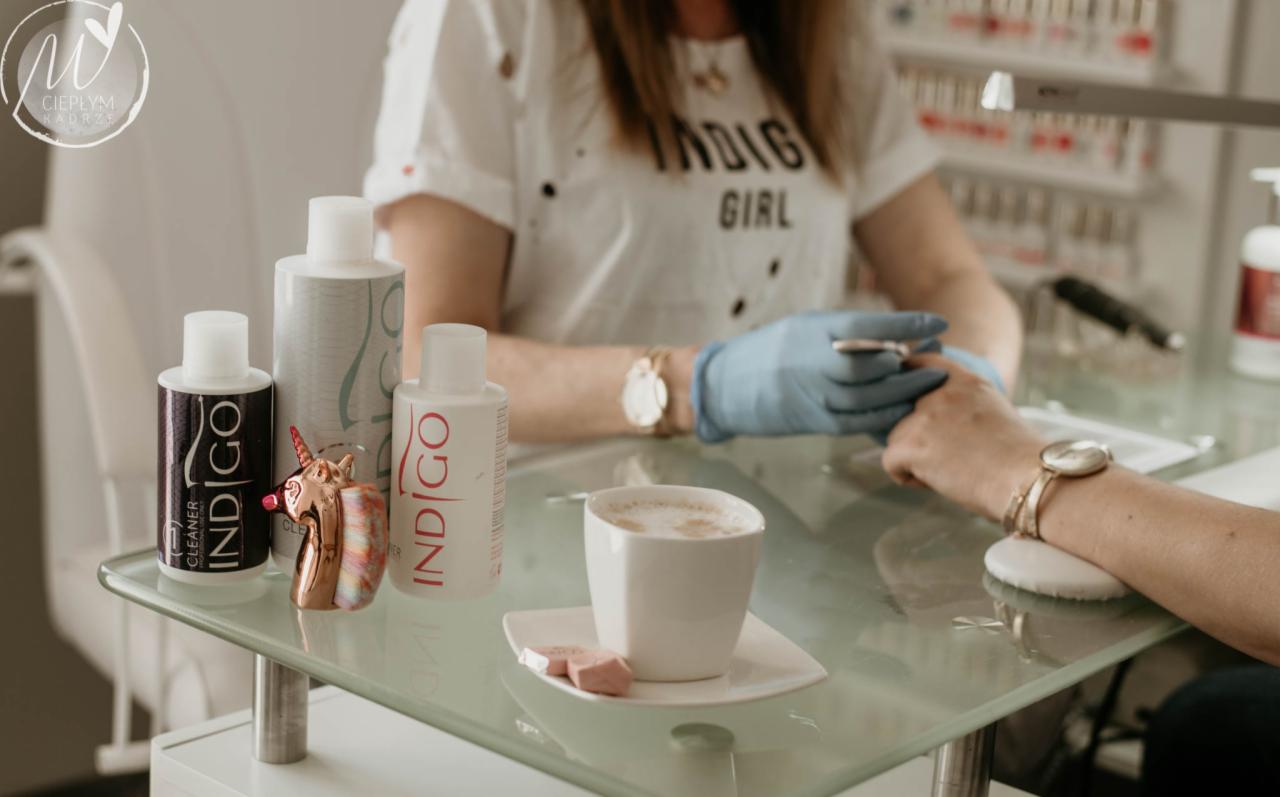 Stylistka paznokci w niebieskich rękawiczkach opracowuje skórki klientki przy szklanym stole z kawą i kosmetykami Indigo, obok stoi błyszcząca figurka jednorożca.