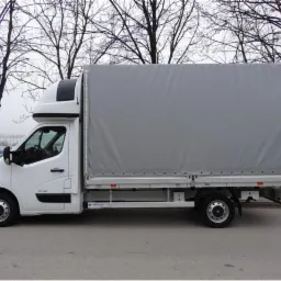 Biały samochód dostawczy z plandeką, widok z boku na tle drzew bez liści i szarej nawierzchni.