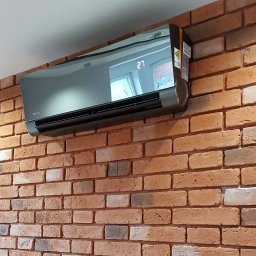 MG KPC Grzegorz Markiewicz - Szary klimatyzator Versu na ceglanej ścianie z widocznym wyświetlaczem temperatury 27 stopni. Nowoczesny design, oświetlenie punktowe.