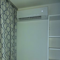 MG KPC Grzegorz Markiewicz - Biały klimatyzator Rotenso na ścianie, obok zasłony w geometryczne wzory i regału. Widoczny wyświetlacz z temperaturą 18 stopni.