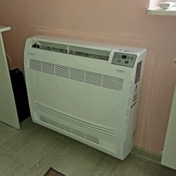 MG KPC Grzegorz Markiewicz - Biały klimatyzator Rotenso Aneru Series, ustawiony przy różowej ścianie, z widocznym panelem sterowania i kratkami wentylacyjnymi. Widoczny fragment podłogi i dywanika.