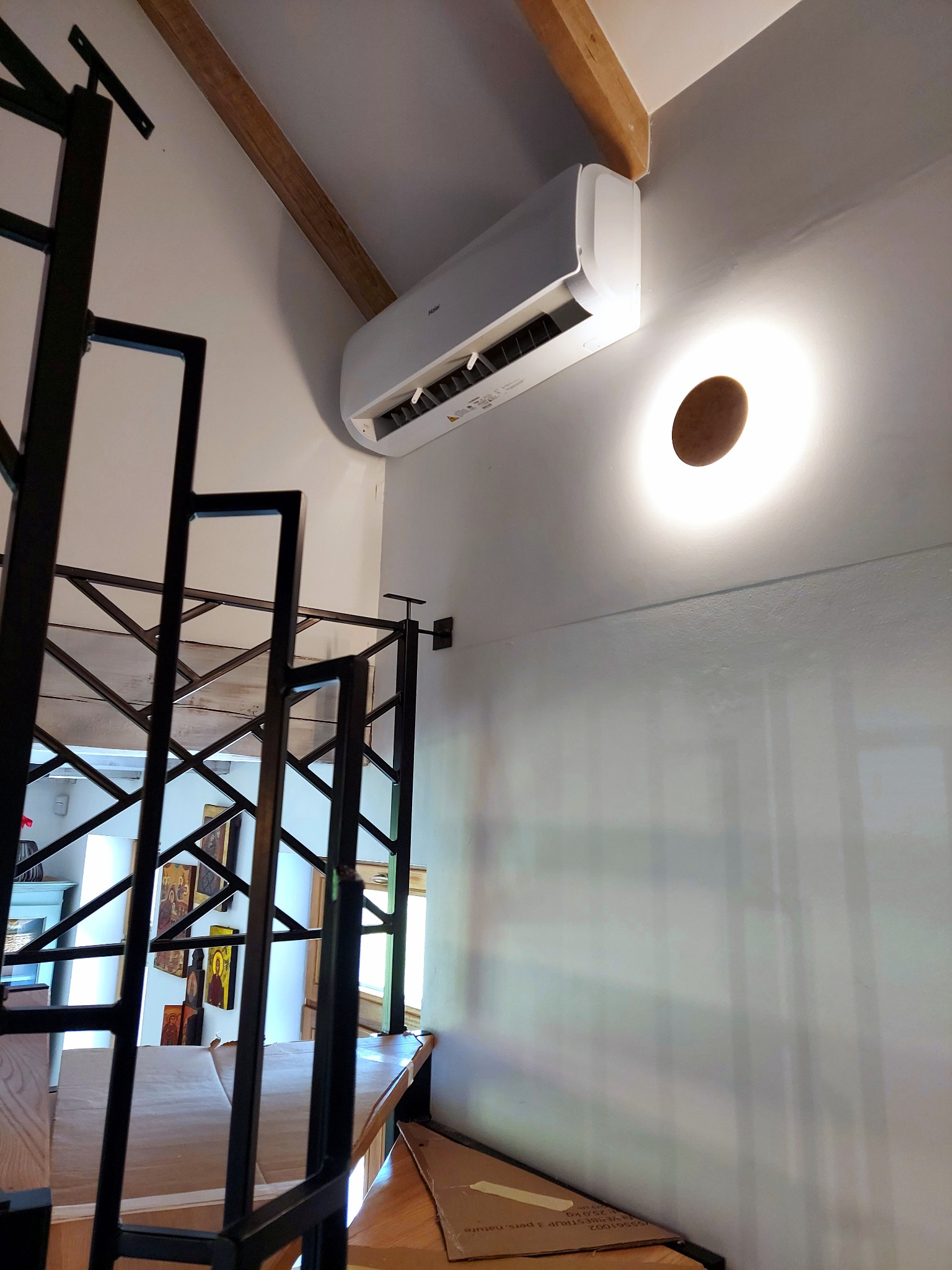 Biały klimatyzator ścienny zamontowany w jasnym wnętrzu, widoczny z dołu, obok nowoczesnej lampy i metalowej konstrukcji schodów.