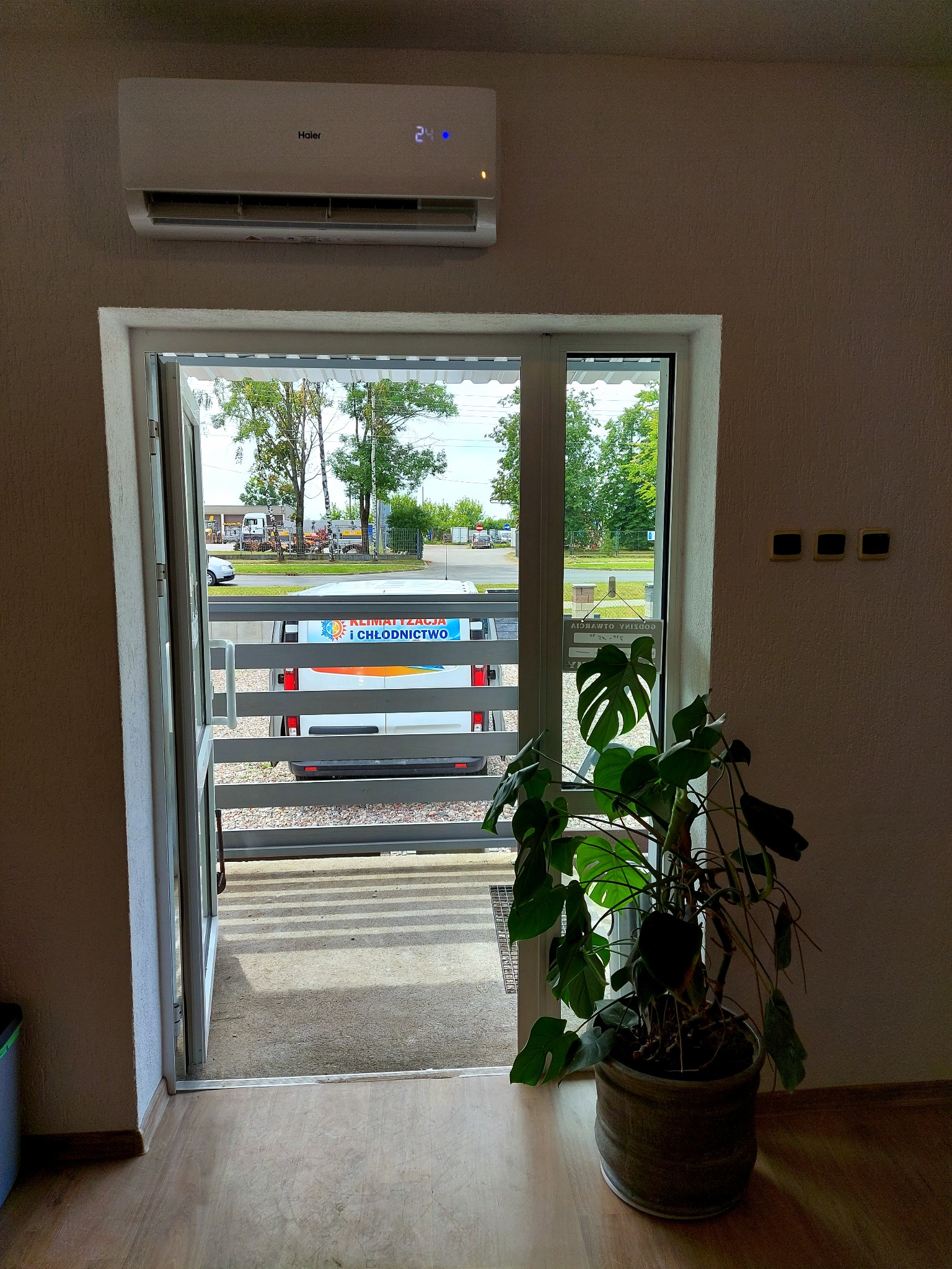 Wnętrze z klimatyzatorem Haier nad otwartymi drzwiami balkonowymi, przez które widać firmowy samochód z napisem 'Klimatyzacja i Chłodnictwo'.