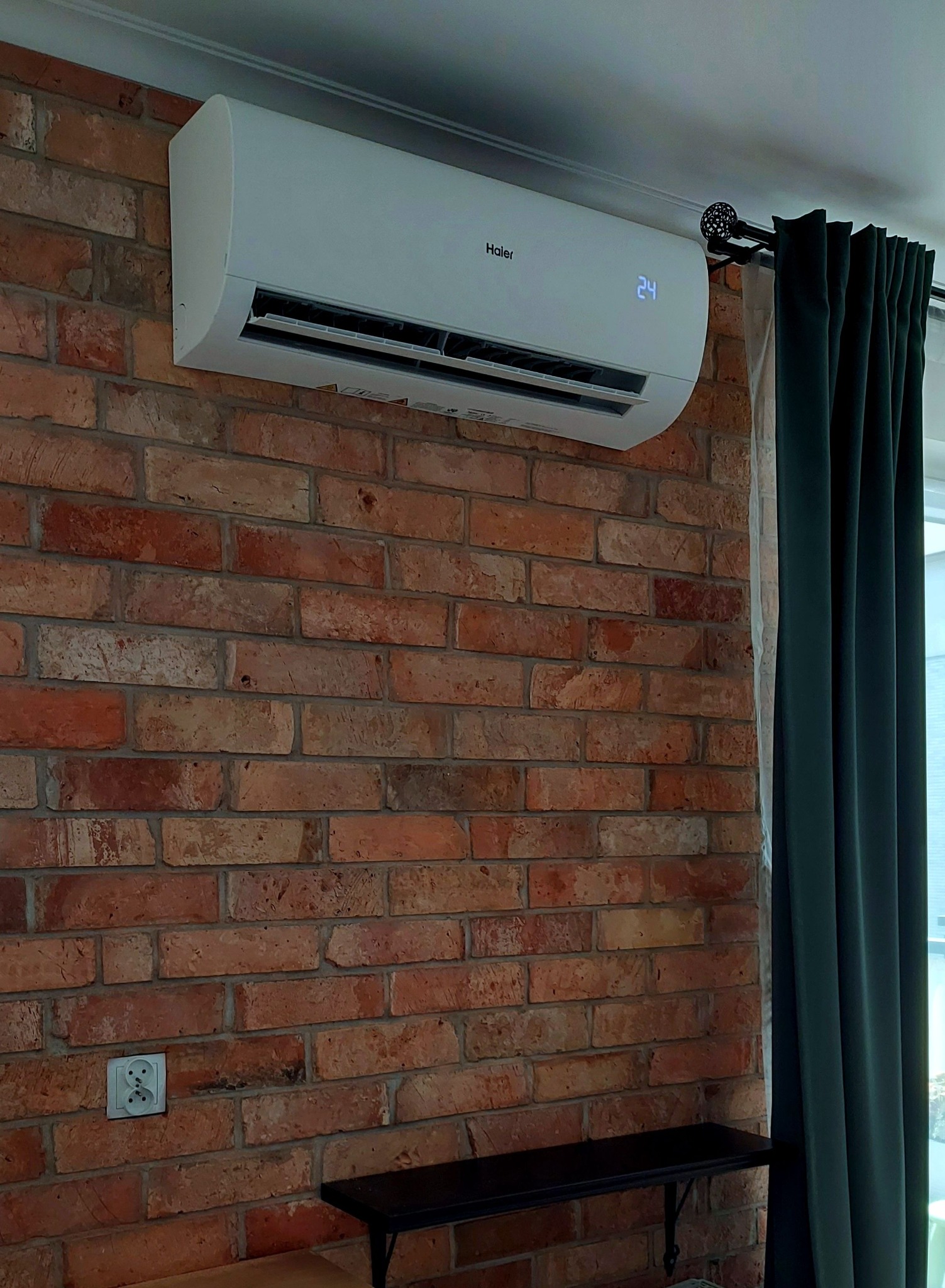 Biały klimatyzator Haier na ceglanej ścianie, z widocznym wyświetlaczem temperatury 24°C, obok ciemnozielonych zasłon i czarnego stolika. Widoczne gniazdko elektryczne.