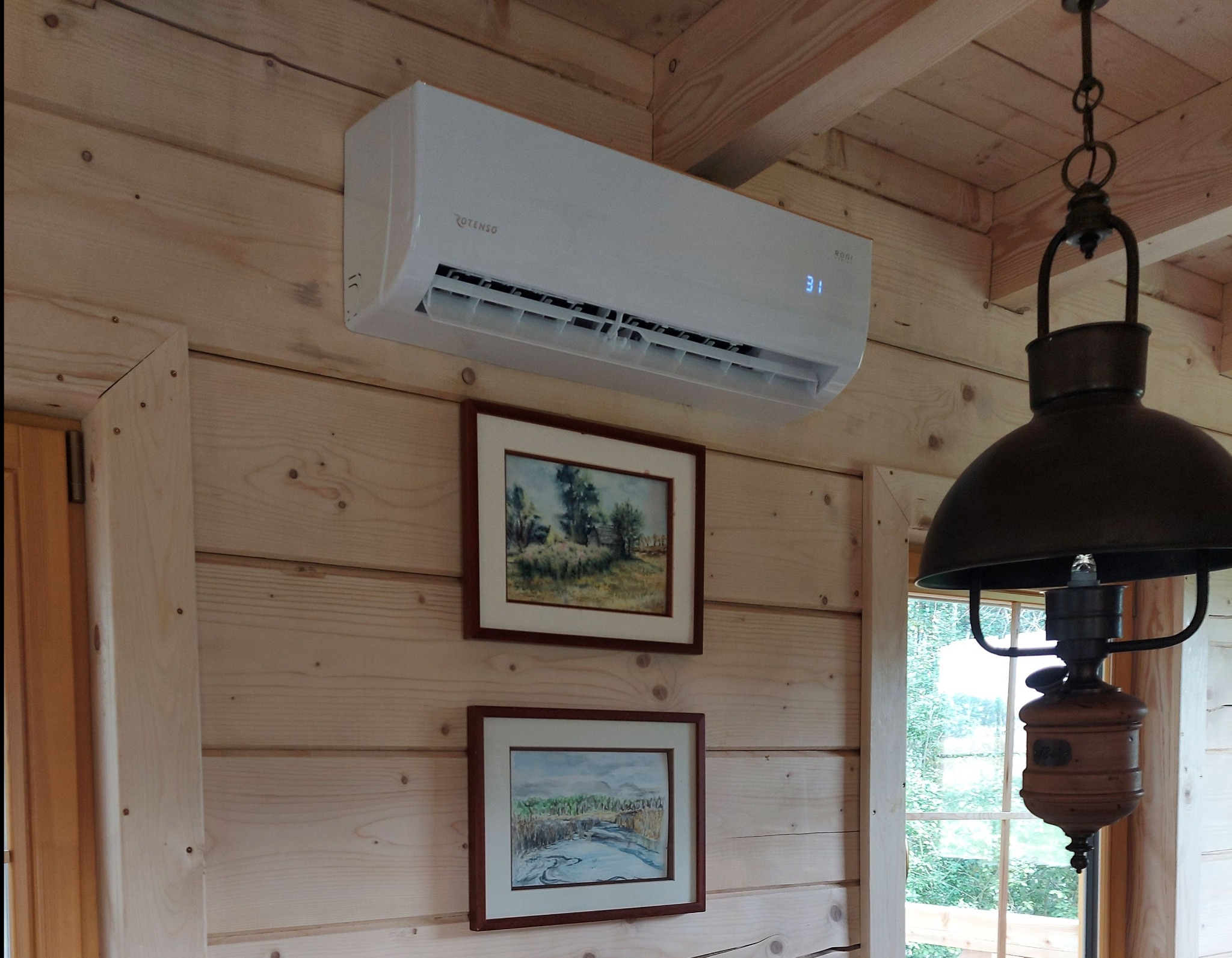 Biały klimatyzator Rotenso na drewnianej ścianie w rustykalnym wnętrzu, pod nim dwa obrazy w ramkach, obok lampa wisząca. Widoczny fragment okna.
