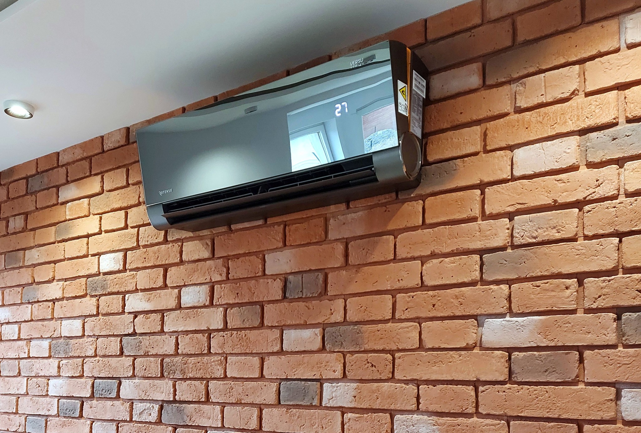 Szary klimatyzator Versu na ceglanej ścianie z widocznym wyświetlaczem temperatury 27 stopni. Nowoczesny design, oświetlenie punktowe.