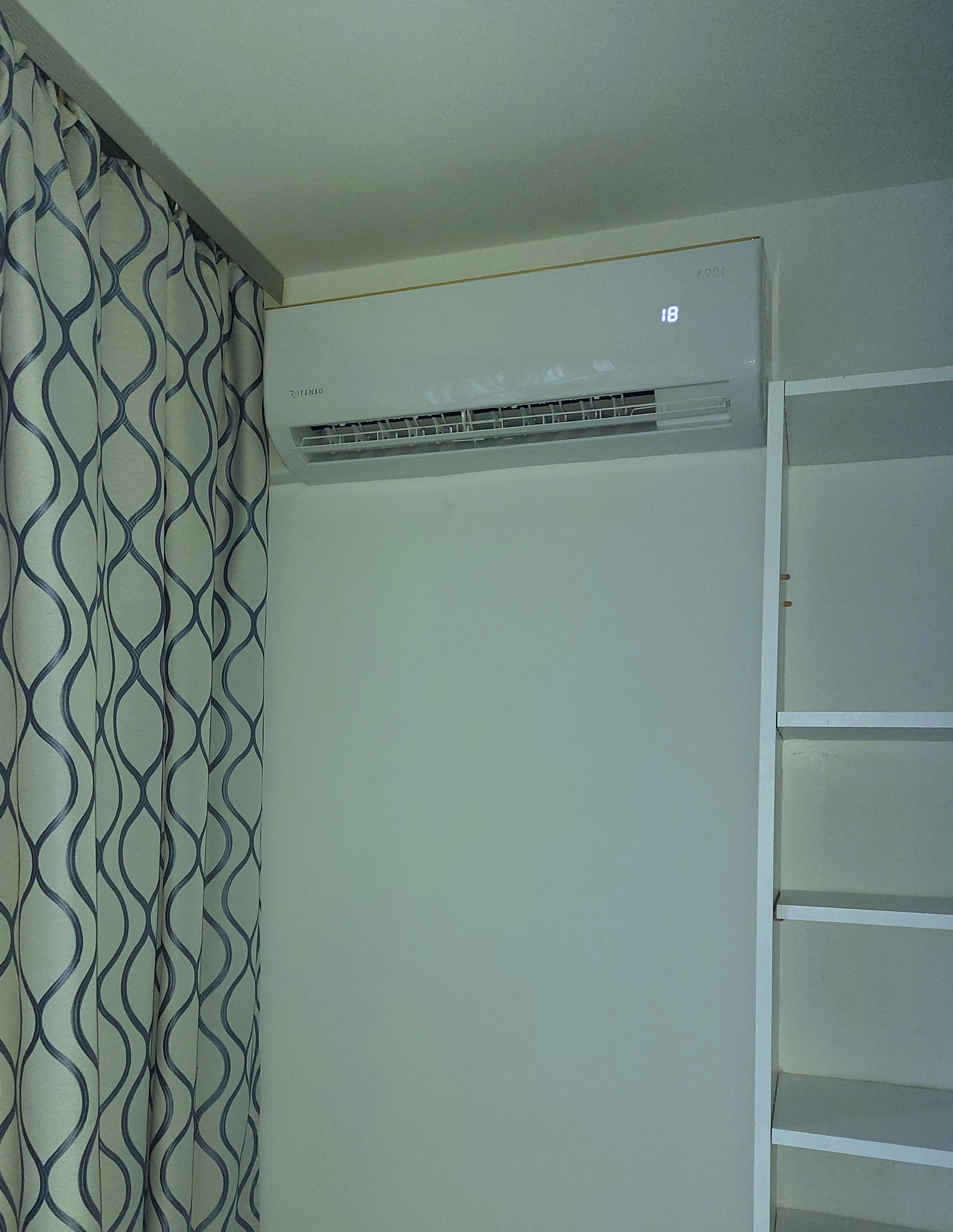 Biały klimatyzator Rotenso na ścianie, obok zasłony w geometryczne wzory i regału. Widoczny wyświetlacz z temperaturą 18 stopni.