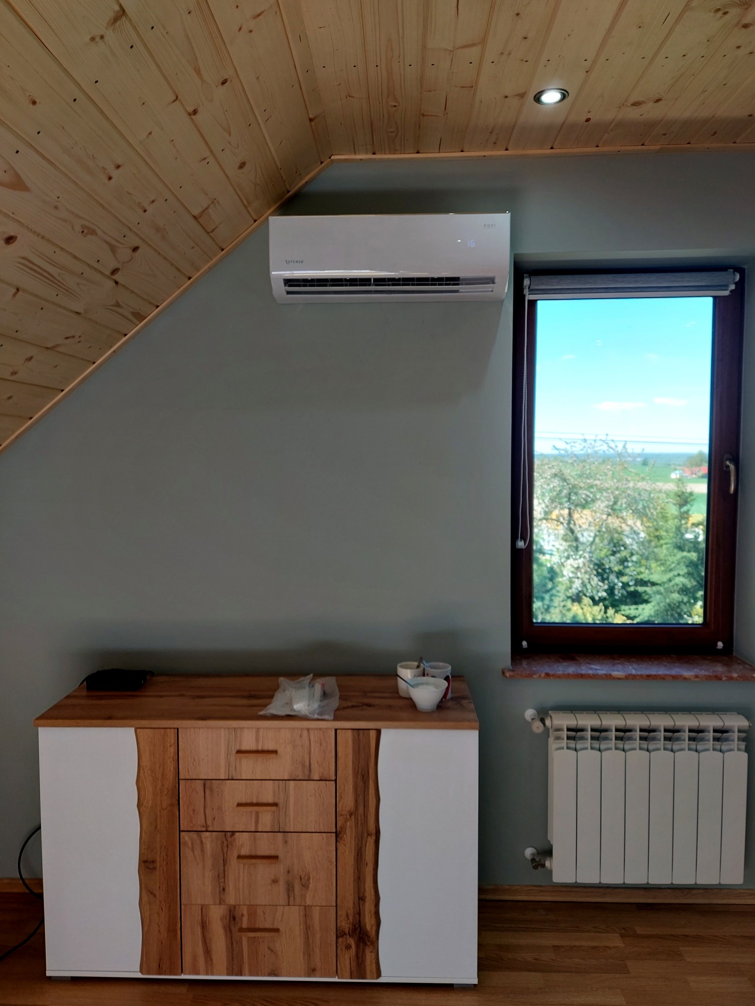 Biały klimatyzator na szarej ścianie w pokoju z drewnianym sufitem i komodą. Widok na zewnątrz przez okno. Grzejnik pod oknem.