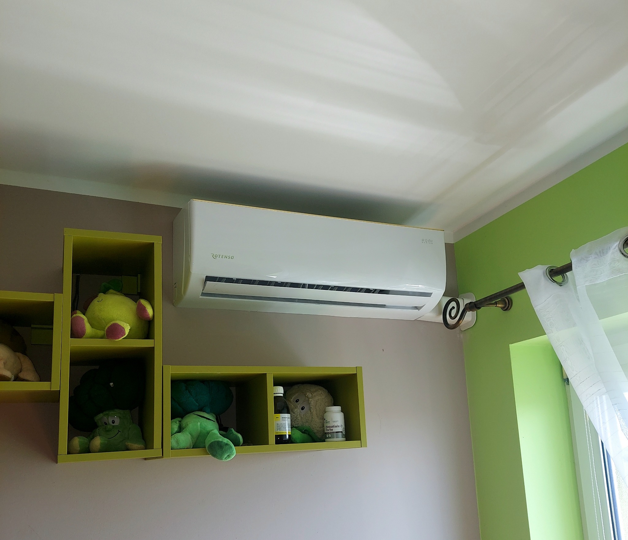 Biała klimatyzacja Rotenso zamontowana na ścianie w pokoju dziecięcym, obok zielonych półek z pluszakami i okna z firanką. Widok z dołu.