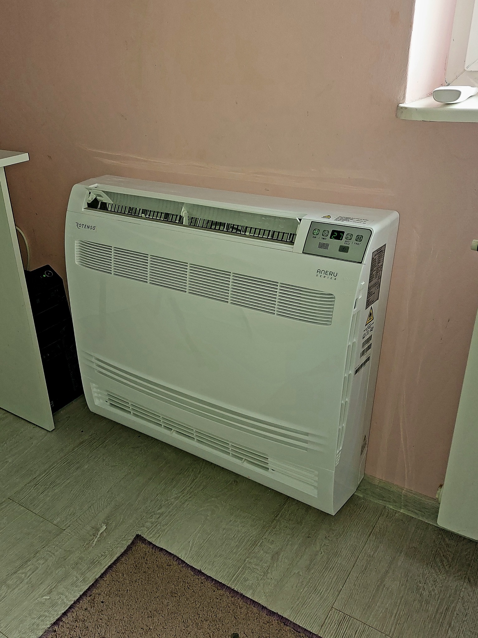 Biały klimatyzator Rotenso Aneru Series, ustawiony przy różowej ścianie, z widocznym panelem sterowania i kratkami wentylacyjnymi. Widoczny fragment podłogi i dywanika.