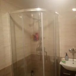 Wykończona łazienka z narożną kabiną prysznicową, beżowe płytki na ścianach i brązowe akcenty, umywalka z szafką w rogu.