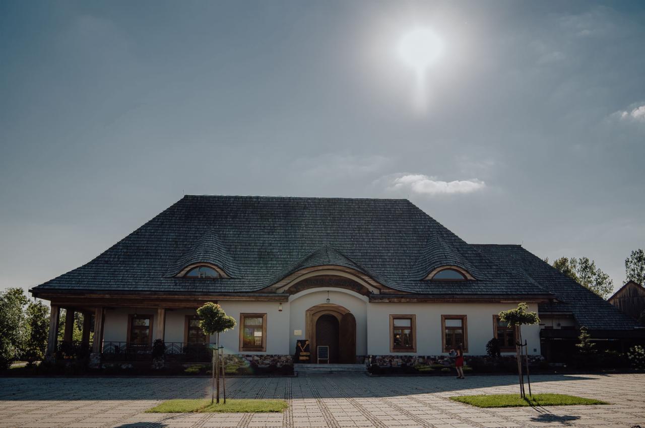 Duży budynek z drewnianym dachem krytym gontem, owalnymi oknami na poddaszu i kolumnami po lewej stronie, oświetlony silnym słońcem.