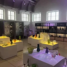 Ekspozycja szkła artystycznego w przestronnym wnętrzu z wysokimi oknami, oświetlona punktowo, prezentująca wazony, misy i inne formy szklane na białych podestach i w przeszklonych witrynach.