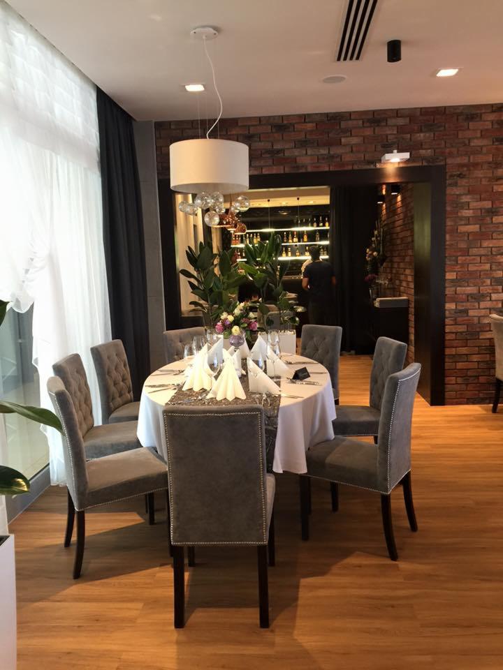 Elegancko nakryty okrągły stół w restauracji z szarymi krzesłami, oświetlony nowoczesną lampą, na tle ceglanej ściany i wejścia do baru.