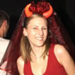 Uśmiechnięta kobieta w czerwonej sukience i welonie z rogami diabła na głowie, prawdopodobnie podczas wieczoru panieńskiego w klubie, z widoczną sylwetką mężczyzny w tle.