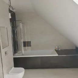 Nowoczesna łazienka na poddaszu z wanną za szklanym parawanem, jasnymi płytkami imitującymi marmur i ciemnoszarą zabudową wanny. Widoczne niedokończone podłączenie elektryczne oświetlenia.