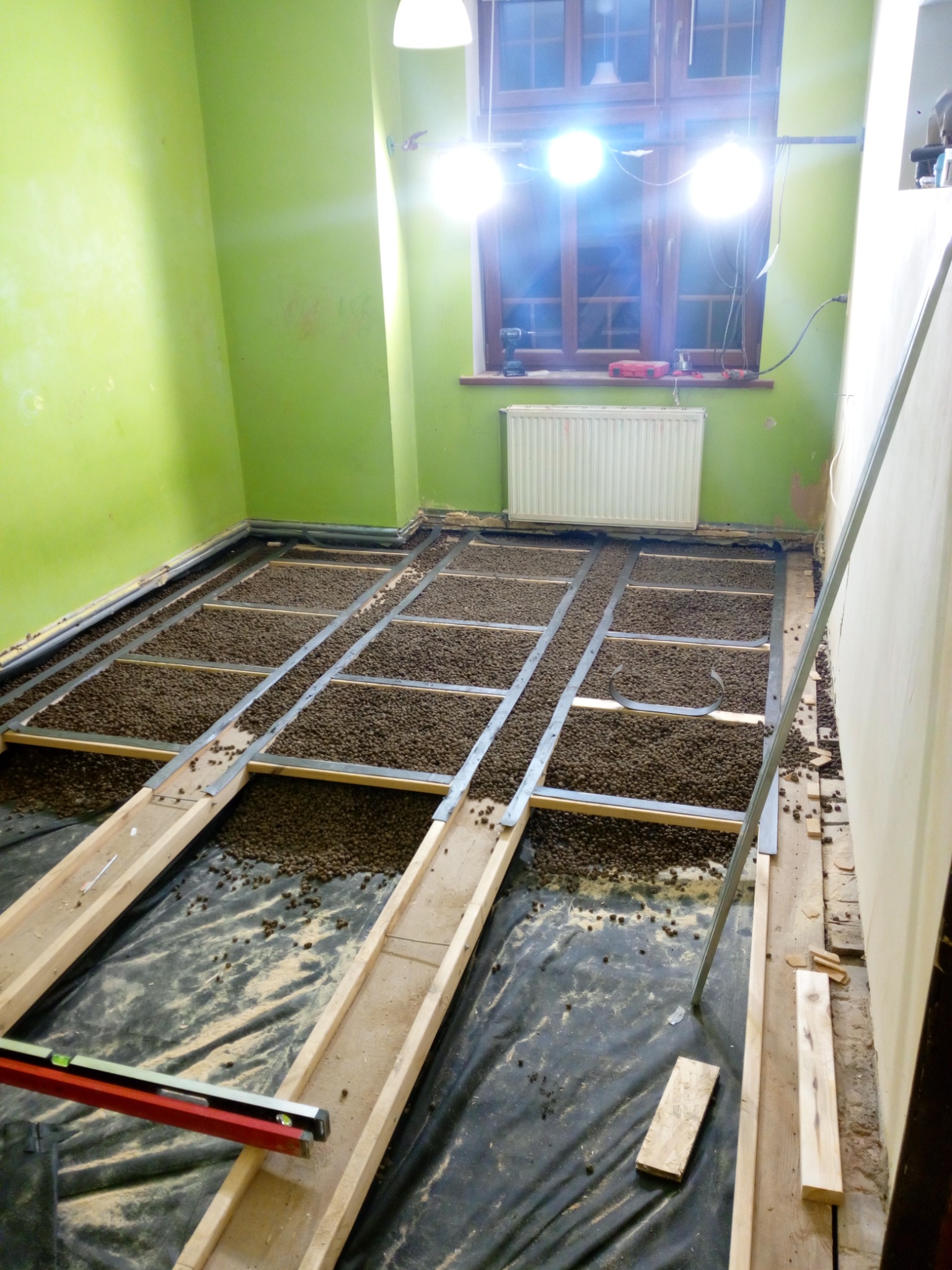 Przygotowanie podłoża pod panele: drewniane legary, wypełnienie granulatem wyrównującym, czarna folia izolacyjna, poziomica leżąca na legarze, ściana w kolorze limonkowym.