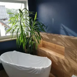 Biała wanna owinięta folią ochronną w trakcie prac, ściana w kolorze granatowym, drewnopodobne płytki na ścianie z wnęką, doniczka z zieloną rośliną przy oknie, podłoga wyłożona panelami.