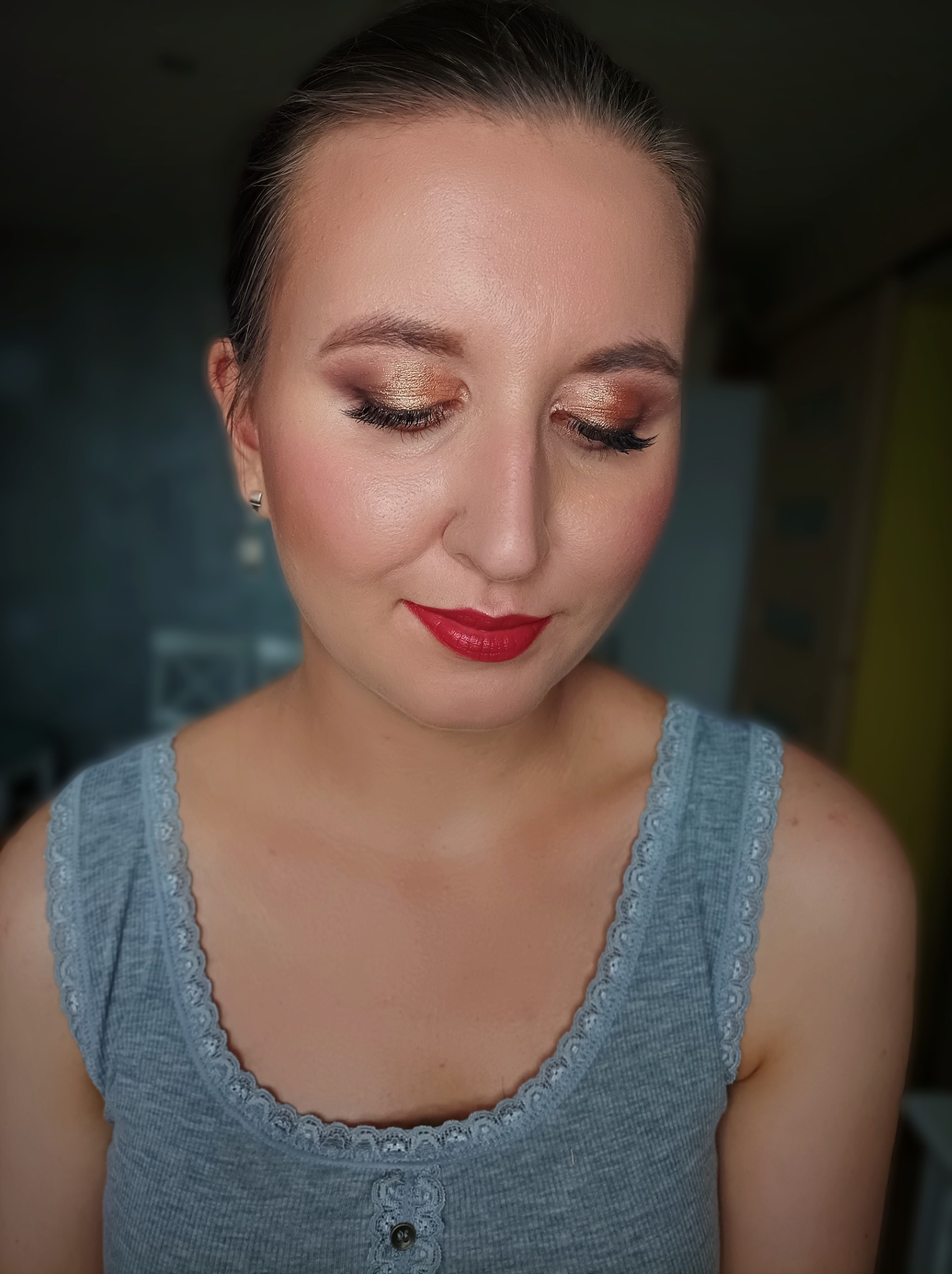 Portret kobiety z zamkniętymi oczami, prezentującej złoto-brązowy makijaż oczu z wyraźnie wytuszowanymi rzęsami i intensywnie czerwoną szminką. Delikatny róż na policzkach i upięte włosy. Ubrana...