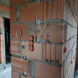 Instalacja elektryczna w surowym budynku: widoczne pomarańczowe puszki pod gniazdka i kable wystające ze ścian z czerwonej cegły.