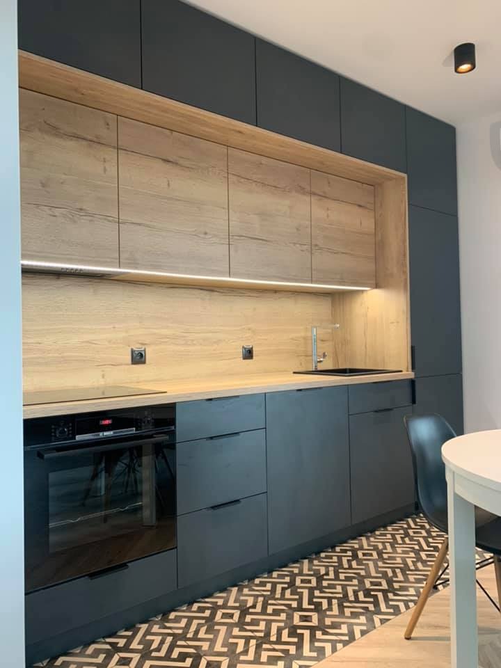 Nowoczesna kuchnia z ciemnoszarymi matowymi frontami szafek i drewnianymi akcentami, podświetlenie LED pod górnymi szafkami, wzorzysta podłoga, czarny zlew i armatura.