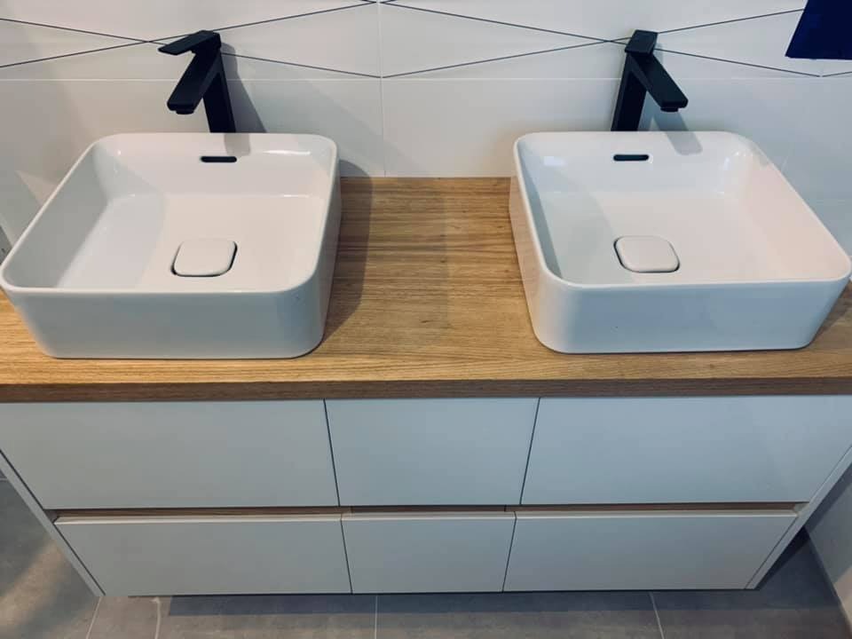 Dwie kwadratowe, białe umywalki nablatowe z czarnymi bateriami, zamontowane na drewnianym blacie nad białymi szafkami z szufladami w nowoczesnej łazience z białymi płytkami z czarnymi akcentami.