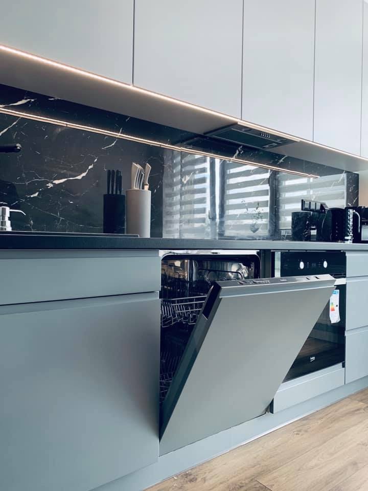 Nowoczesna kuchnia w odcieniach szarości z marmurowym backsplash, podświetleniem LED, otwartą zmywarką i drewnianą podłogą.