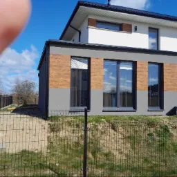 Dwukondygnacyjny dom z elewacją w kolorze białym i szarym z drewnopodobnymi panelami, widoczny zza czarnego ogrodzenia siatkowego na tle błękitnego nieba z chmurami, z widocznym palcem w lewym...