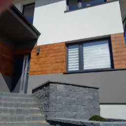 Fragment elewacji domu po remoncie: biała góra, szara środkowa część i imitacja drewnianej okładziny na dole, szare okna z żaluzjami, wejście z kamiennymi schodami i murkiem.