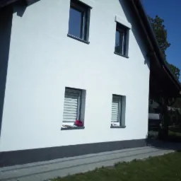 Boczny widok domu z białą elewacją i szarym cokołem, z widocznymi czterema oknami w ciemnych ramach i podcieniem z ciemnym dachem, na parapecie jednego z okien leży czerwony przedmiot.