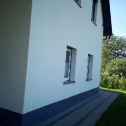 Wykończona elewacja domu jednorodzinnego z białym tynkiem, ciemnoszarym cokołem i szarą kostką brukową, widok z boku na tle zielonego ogrodu.