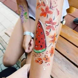 Malunek na ramieniu dziecka przedstawiający plaster arbuza i pomarańczowe kwiaty, na drugim ramieniu fioletowe kwiaty.