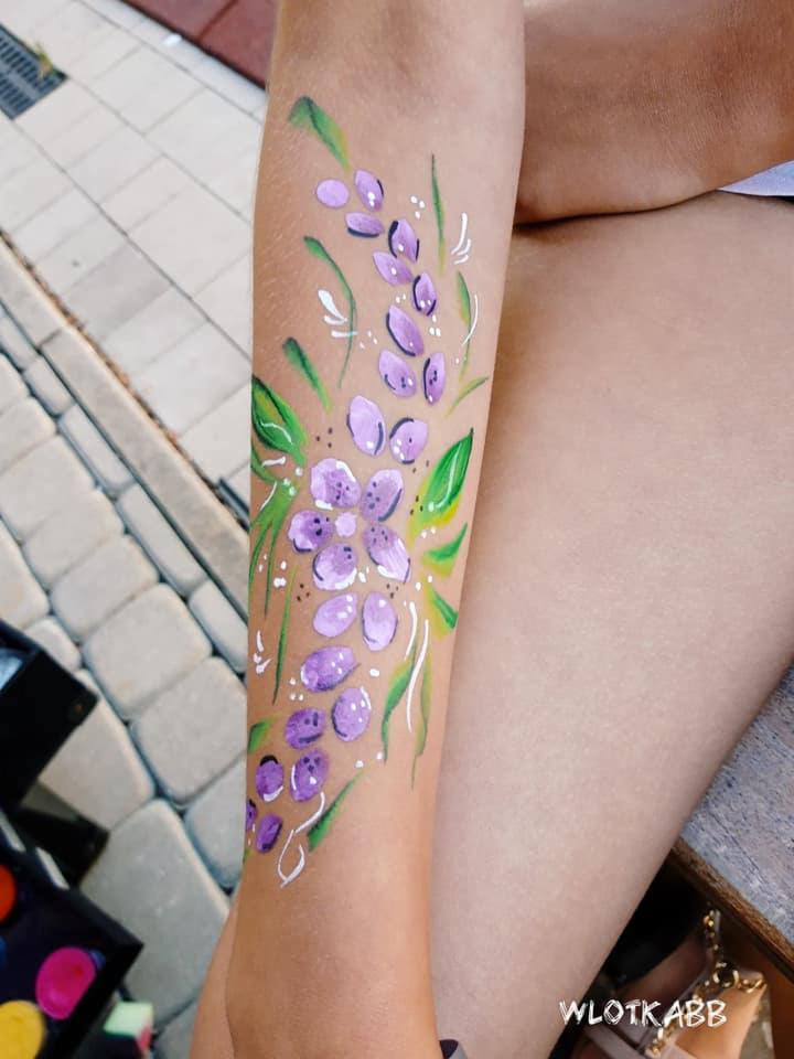 Ręcznie malowany motyw kwiatowy w odcieniach fioletu i zieleni na ramieniu, wykonany w Bielsko-Białej. Widoczny fragment palety z farbami.