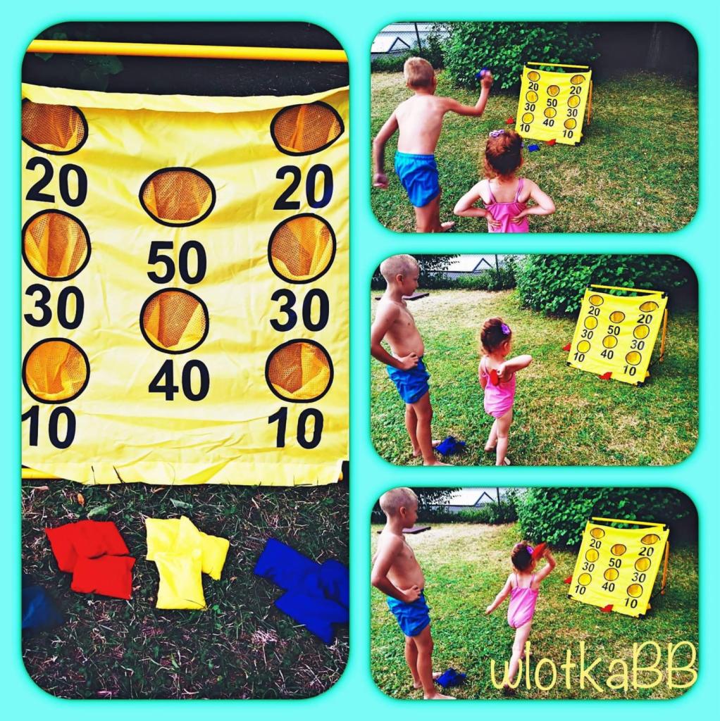 Dzieci grają w rzut woreczkami do tarczy punktowej na trawie; żółta tarcza z otworami oznaczonymi punktami 10-50, obok leżą kolorowe woreczki.